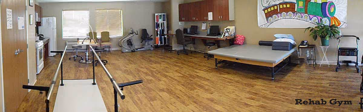 A rehabilitation gym room with wooden flooring, parallel bars for walking exercises, exercise equipment including a stationary bike, chairs, a treatment table with pillows, cabinets, a desk, and a colorful train mural on the wall.