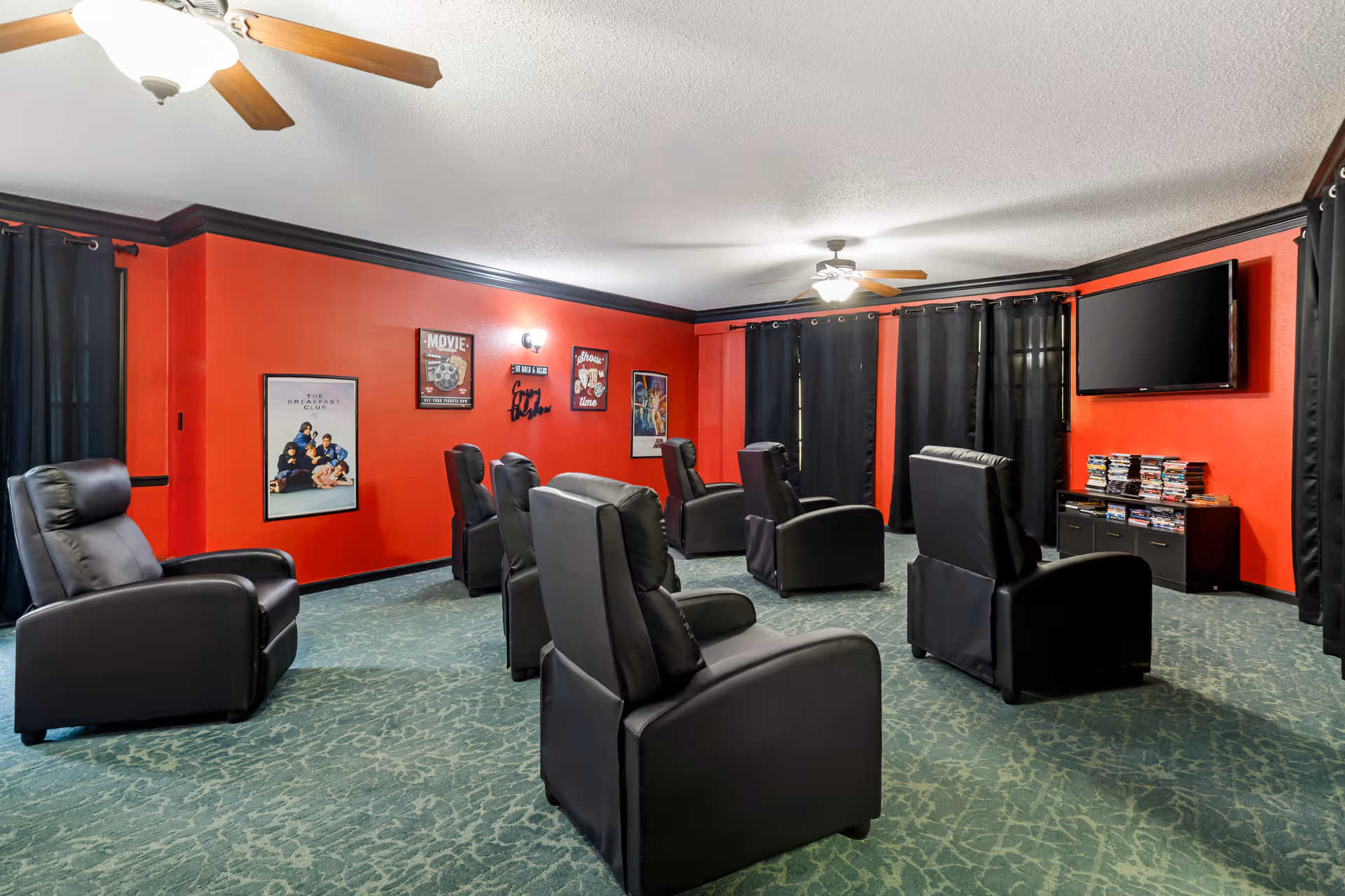 Red-walled senior living theater with black recliners, curtains, and a wall-mounted TV.