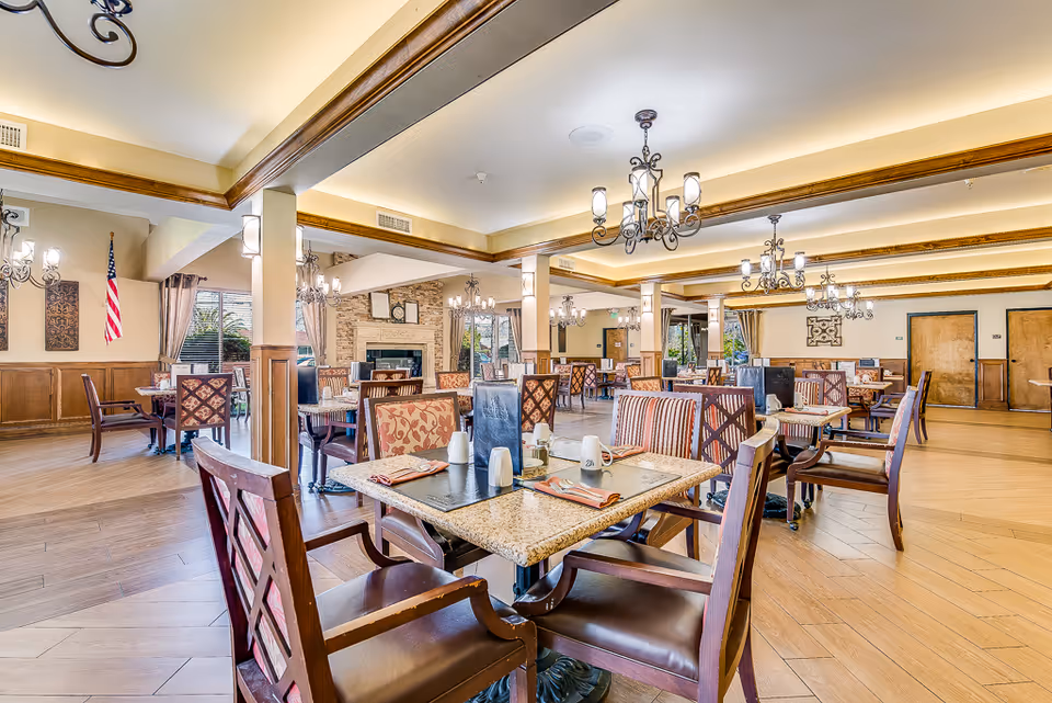 A spacious dining room in a senior living facility with multiple tables and chairs arranged neatly. The room features wooden flooring, decorative chandeliers hanging from the ceiling, and large windows with curtains allowing natural light. There is a fireplace with a clock above it and an American flag displayed on one wall.