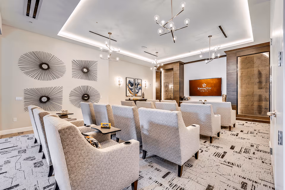 A modern, well-lit room with rows of comfortable beige armchairs facing a large wall-mounted screen displaying the MorningStar Assisted Living & Memory Care logo. The room features contemporary light fixtures, abstract wall art, and a patterned carpet, creating a cozy and inviting atmosphere.