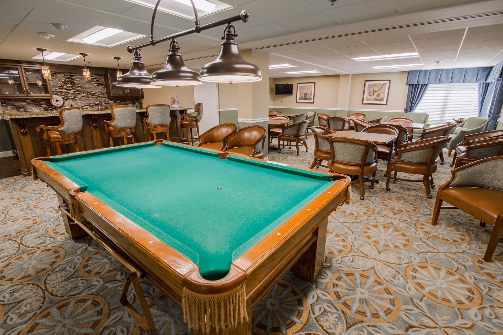 A recreational room featuring a green felt pool table in the foreground, surrounded by several brown leather and fabric chairs. In the background, there is a bar area with high chairs and a mosaic tile backsplash. The room has patterned carpet flooring, framed artwork on the walls, a wall-mounted TV, and large windows with blue curtains allowing natural light.