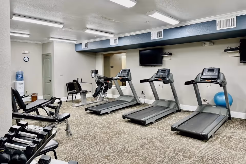Interior view of a fitness room in a senior living facility with three treadmills, exercise bikes, a set of dumbbells, and a water cooler. The room has beige walls, carpeted floor, and two wall-mounted televisions.