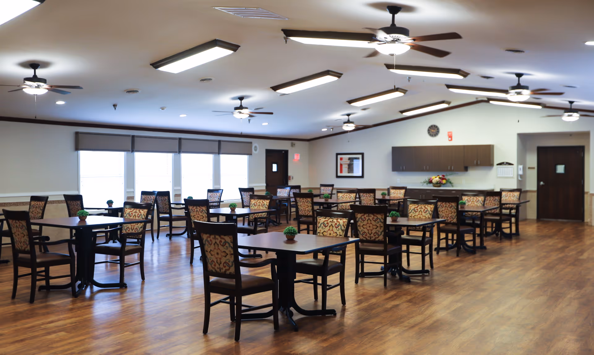 A spacious dining room with multiple square tables and patterned chairs arranged neatly on a wooden floor. The room has ceiling fans with lights, large windows with blinds, and a wall with cabinets and a clock. Small potted plants are placed on each table.