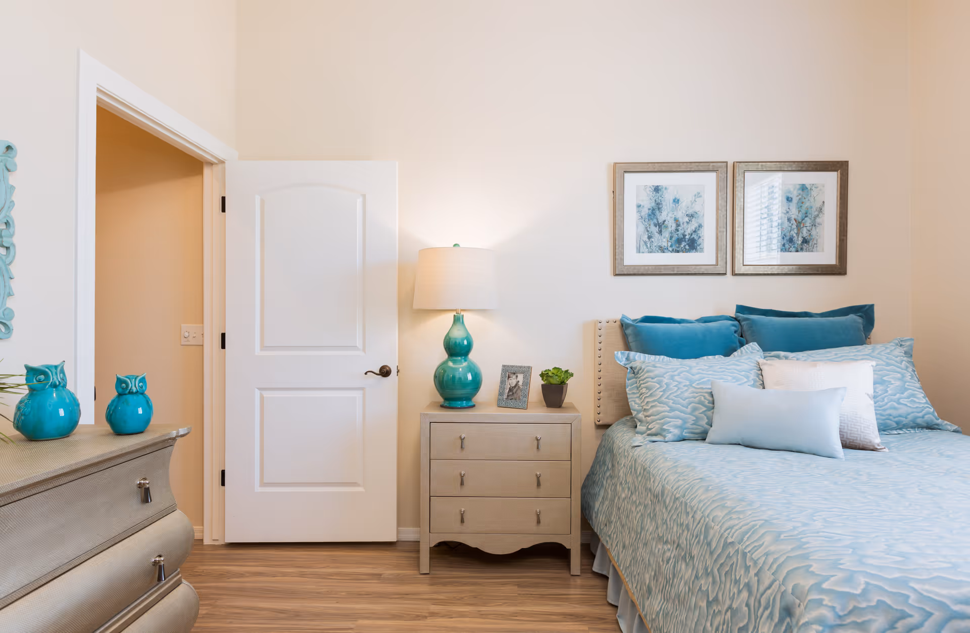 A cozy bedroom with a neatly made bed featuring multiple blue and white pillows and a light blue patterned bedspread. Next to the bed is a wooden nightstand with a turquoise lamp, a small framed photo, and a small potted plant. On the left side, there is a dresser with two turquoise owl figurines and a decorative mirror on the wall. The room has light-colored walls and wooden flooring, with an open white door leading to another room.