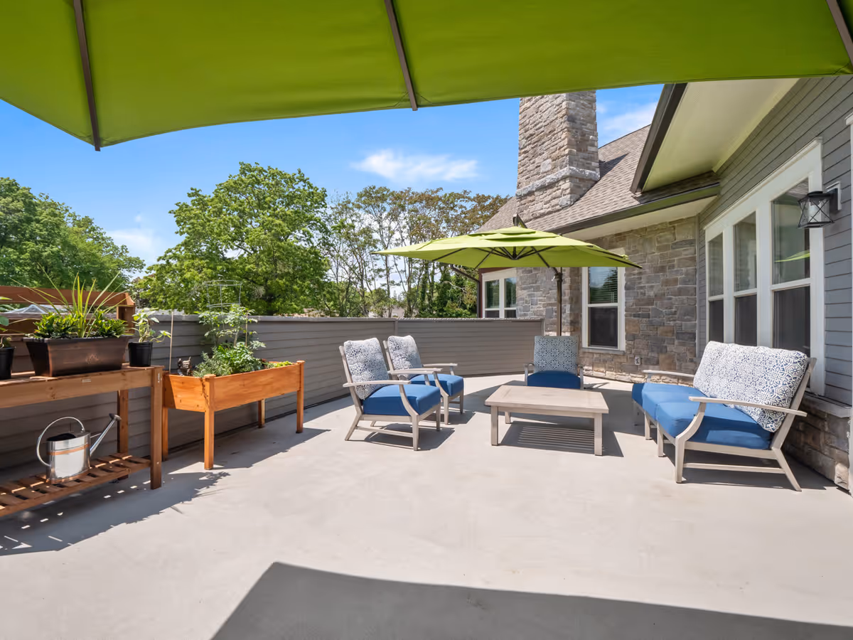 Outdoor patio area with blue cushioned seating including a sofa and two chairs around a low table, shaded by green umbrellas. There are raised wooden planters with plants and a watering can on a shelf. The patio is adjacent to a stone and gray siding building under a clear blue sky.