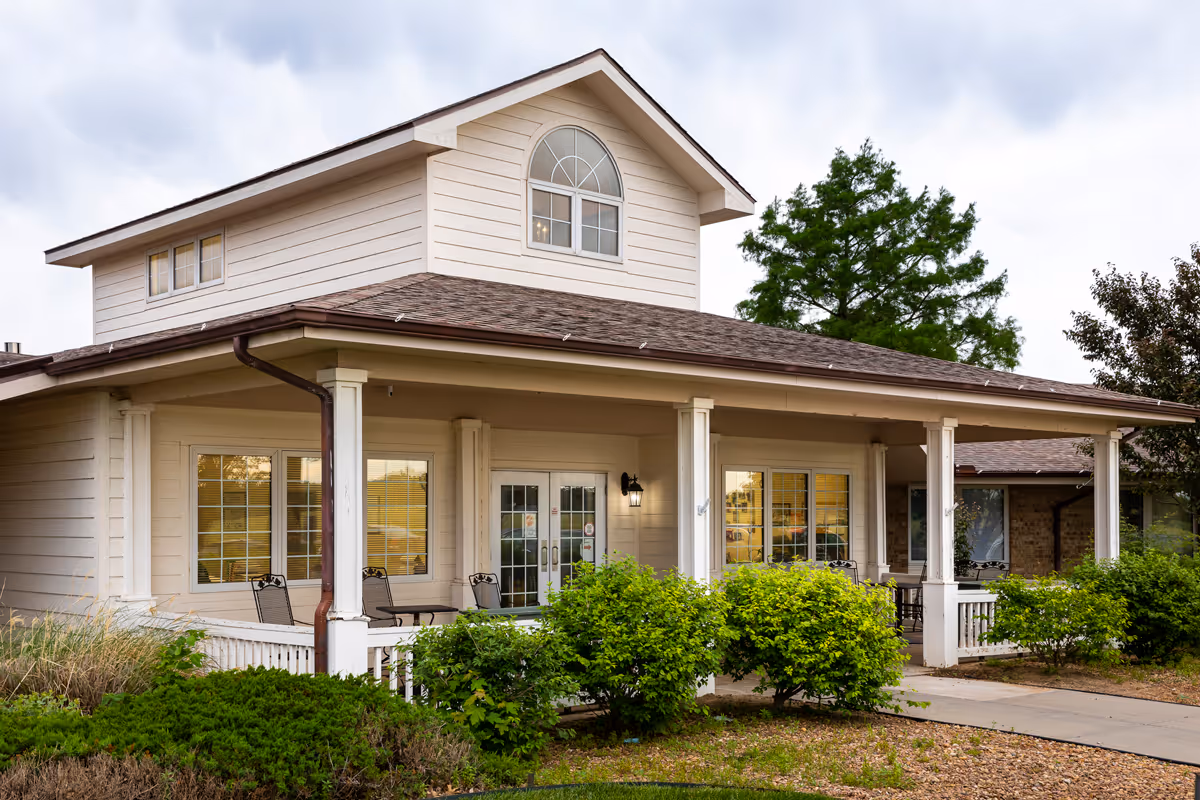 Exterior view of a single-story building with beige siding and a brown shingled roof. The building has a covered porch with white columns and several black metal chairs and tables. There are green bushes and plants in front of the porch, and trees in the background under a cloudy sky.