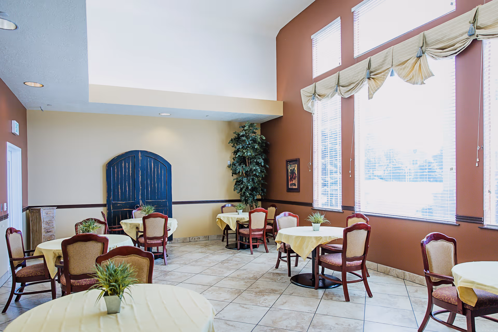 A bright dining room with several round tables covered with yellow tablecloths and small potted plants as centerpieces. The room has large windows with blinds and decorative valances, beige and brown walls, tiled floor, and wooden chairs with cushioned seats and backs. There is a tall artificial plant in the corner and a decorative wooden panel against the far wall.