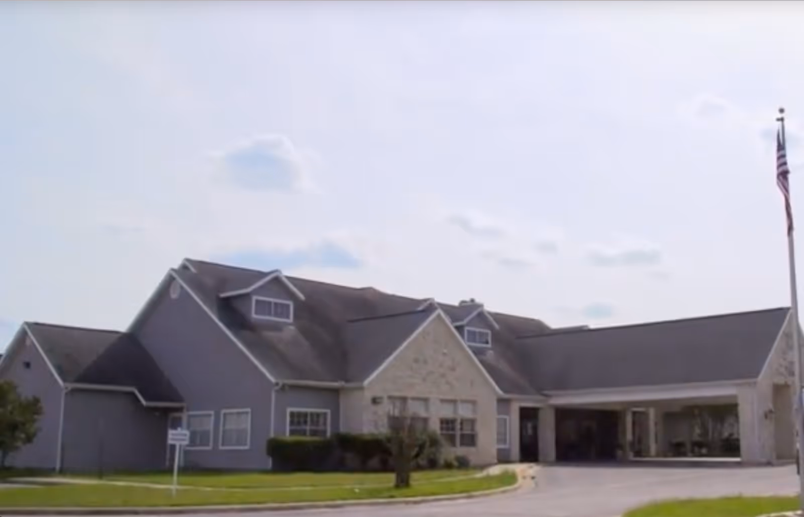 Front exterior of a suburban senior living facility with pitched roofs, a covered entrance/porte-cochère, and an American flag.