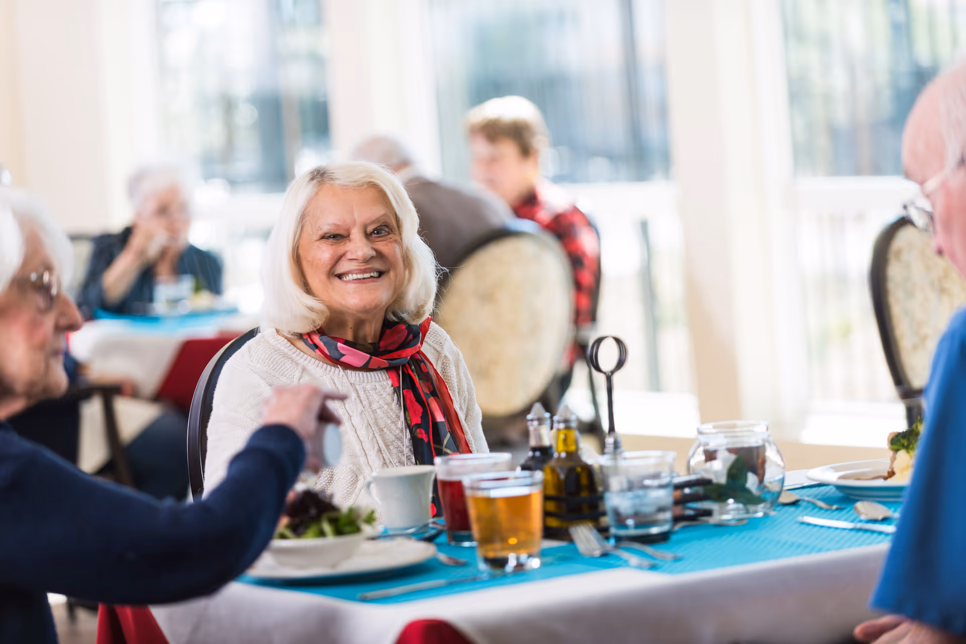 A group of older adults dining in a bright dining room with a smiling woman in the foreground.