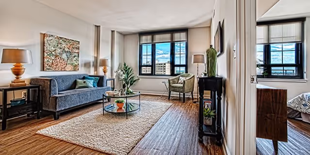 A bright living room with a large window showing a city view. The room features a gray sofa with cushions, two side tables with lamps, a round glass coffee table with plants, a beige rug, a green armchair, and a small black console table with decorative items. The floor is wooden, and there is an adjacent room visible through an open door on the right.