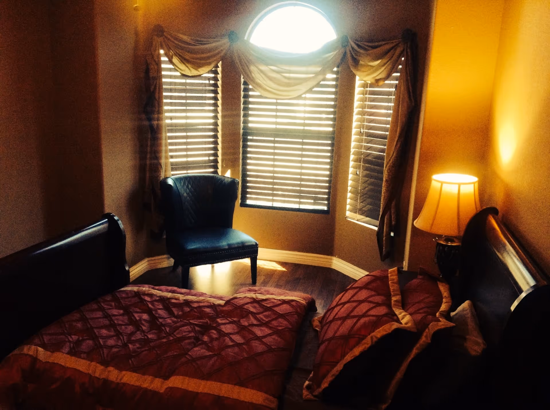 A bedroom with a dark wooden bed covered in a red and gold quilt and pillows. There is a small table lamp on a nightstand next to the bed. A blue upholstered chair is placed near a set of three windows with wooden blinds and decorative drapes. The room has warm lighting and wooden flooring.