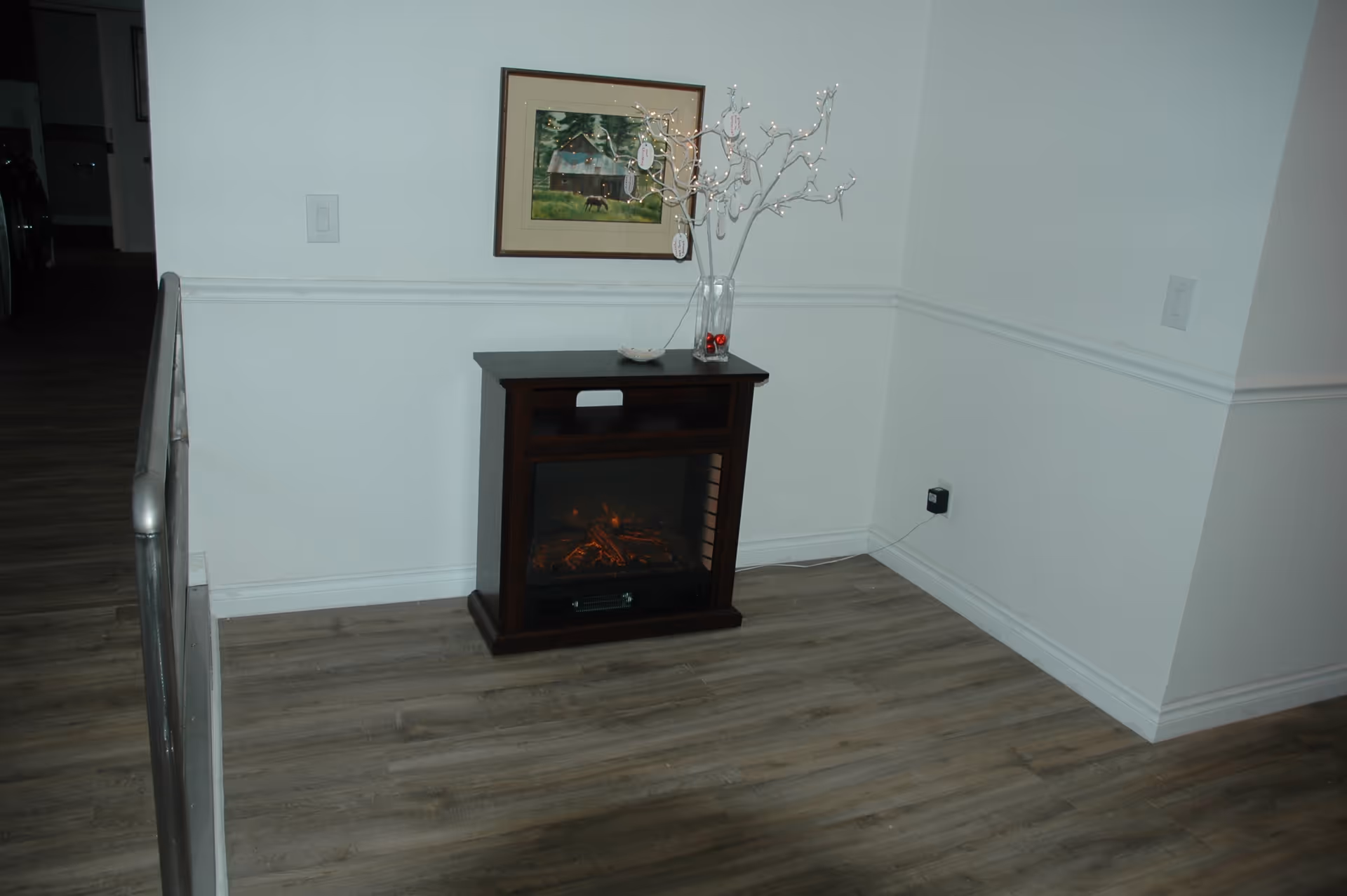 A corner of a room with light-colored walls and wood flooring. There is a small dark wooden electric fireplace with a glowing fire effect. On top of the fireplace is a clear vase holding a decorative white branch with small lights and hanging tags. Above the fireplace, a framed picture of a rustic barn and trees is mounted on the wall. A metal handrail is visible on the left side of the image.