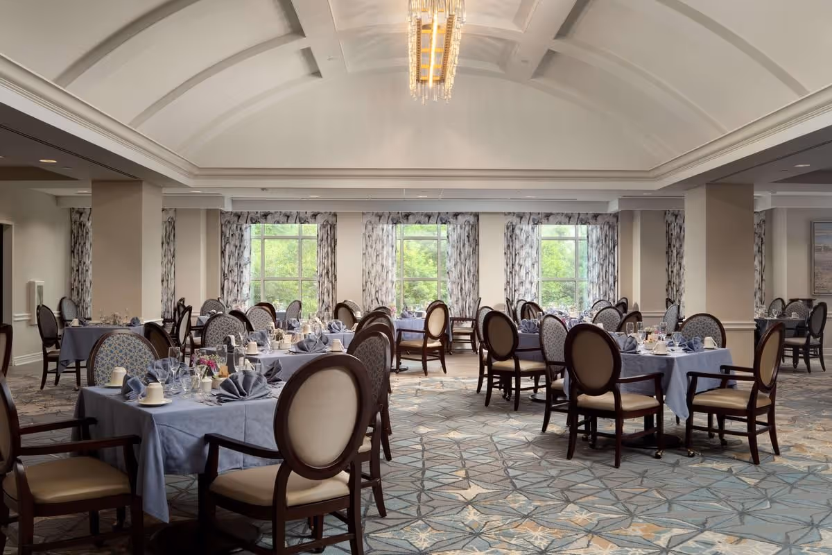 A spacious, elegantly decorated dining room with multiple tables covered in light blue tablecloths. Each table is set with folded napkins, cups, glasses, and small flower arrangements. The room features large windows with patterned curtains allowing natural light to brighten the space. The ceiling has a decorative design with a chandelier hanging in the center. Chairs with cushioned seats and backs surround the tables.