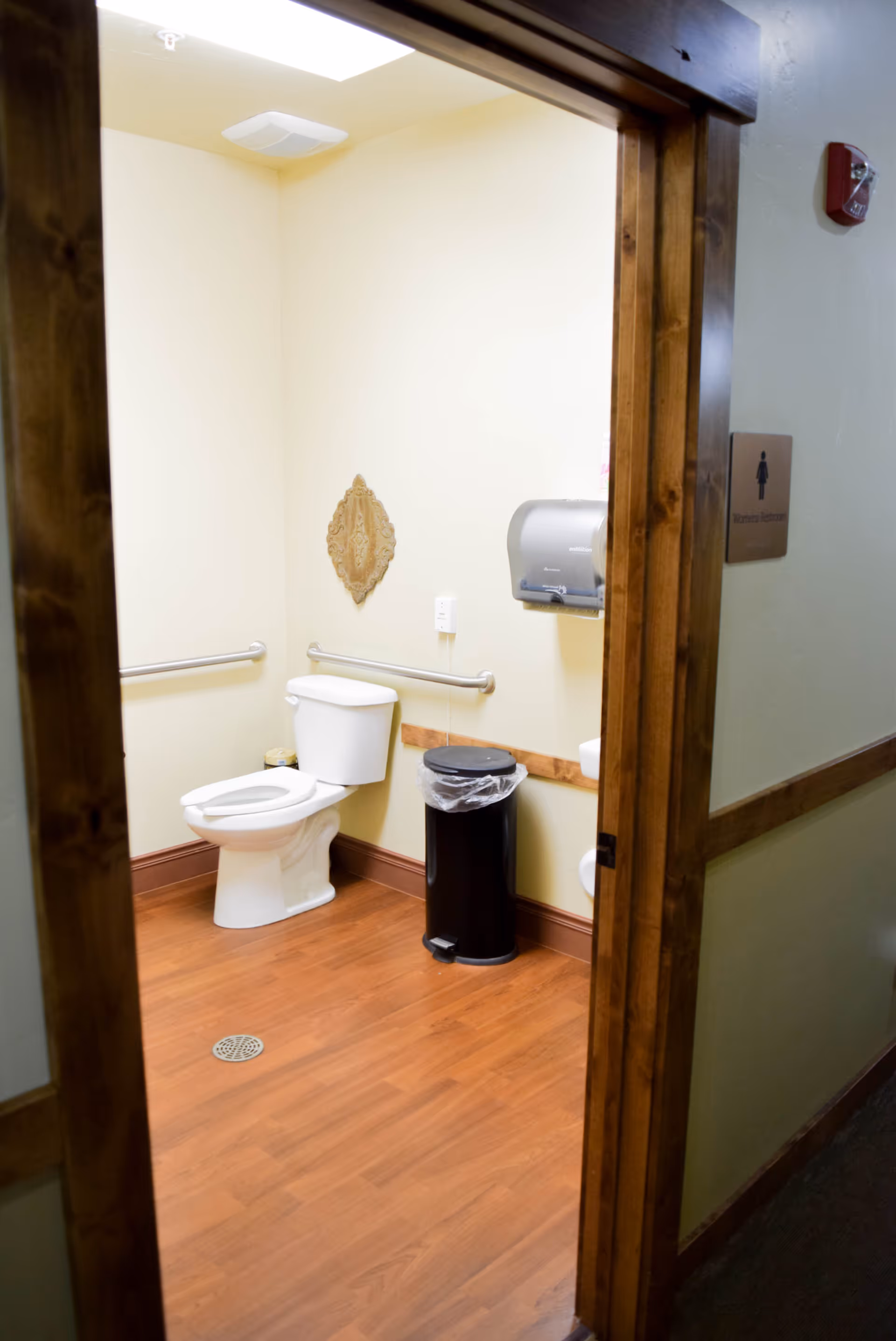Accessible restroom with a toilet, grab bars, wall-mounted paper dispenser and trash can seen through a wood-trimmed doorway.
