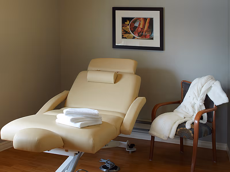 A beige reclining chair with a headrest and folded white towels placed on it, next to a wooden armchair draped with a white robe, set against a gray wall with a framed picture hanging above.