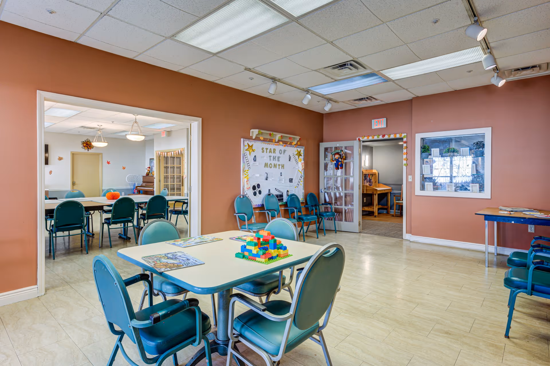 Interior view of a community room with several tables and green chairs arranged around them. One table in the foreground has colorful building blocks and puzzles on it. The walls are painted a warm brown color, and there is a bulletin board labeled 'Star of the Month' with decorations. Through an open doorway, another room with more tables and chairs is visible. The ceiling has fluorescent lighting and track lights.
