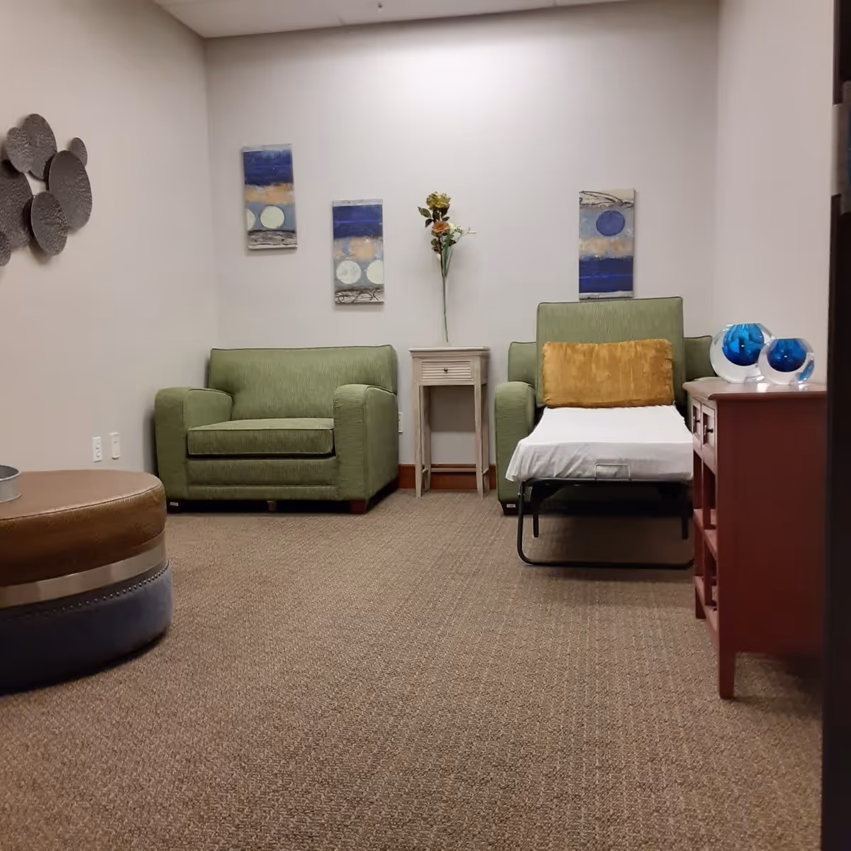 A cozy interior room with two green armchairs, one of which has a fold-out bed extended with a mustard yellow pillow. Between the chairs is a small white side table with a vase holding artificial flowers. On the wall behind the chairs are three vertical abstract paintings with blue, beige, and white tones. To the right is a red wooden cabinet with two blue glass decorative items on top. On the left side of the image is a round ottoman with brown and blue sections, and a metallic wall decoration with circular shapes is mounted on the left wall. The room has beige carpet and white walls.