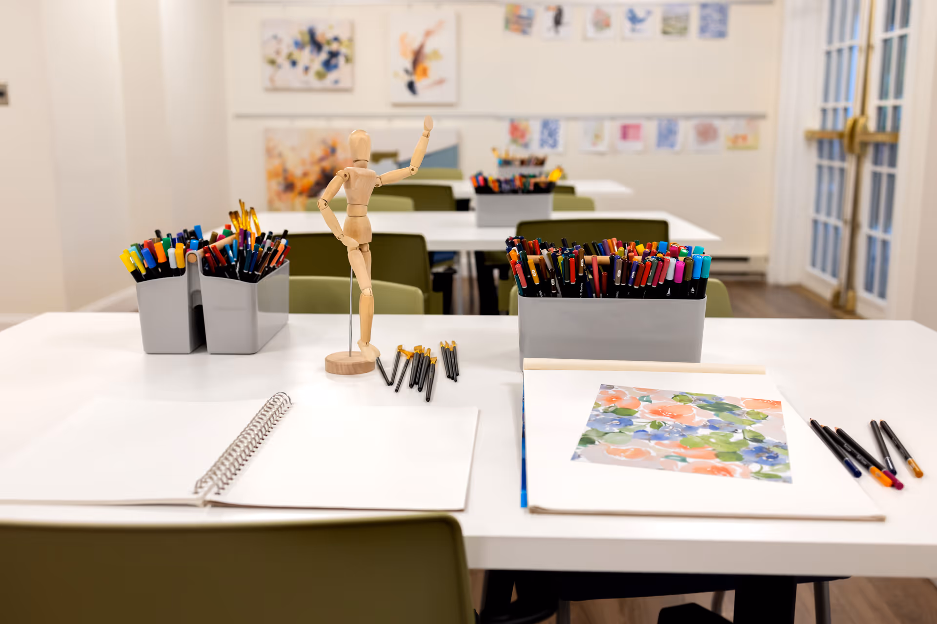 Bright activity room with tables arranged for art projects, pens, markers, sketchbooks and a wooden artist mannequin on the foreground table.