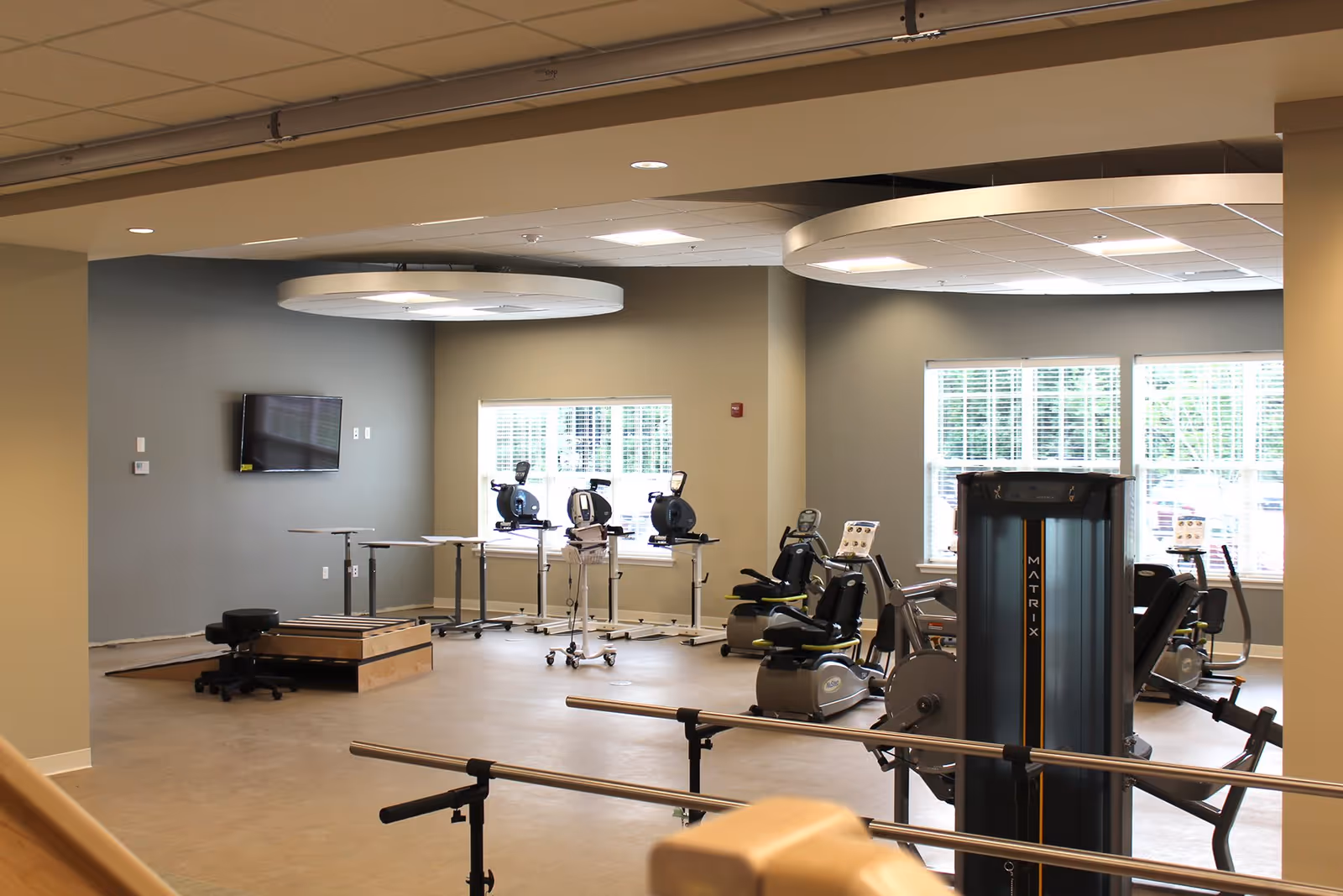 Bright rehabilitation-style fitness room with exercise machines, stationary bikes, parallel bars and large windows.