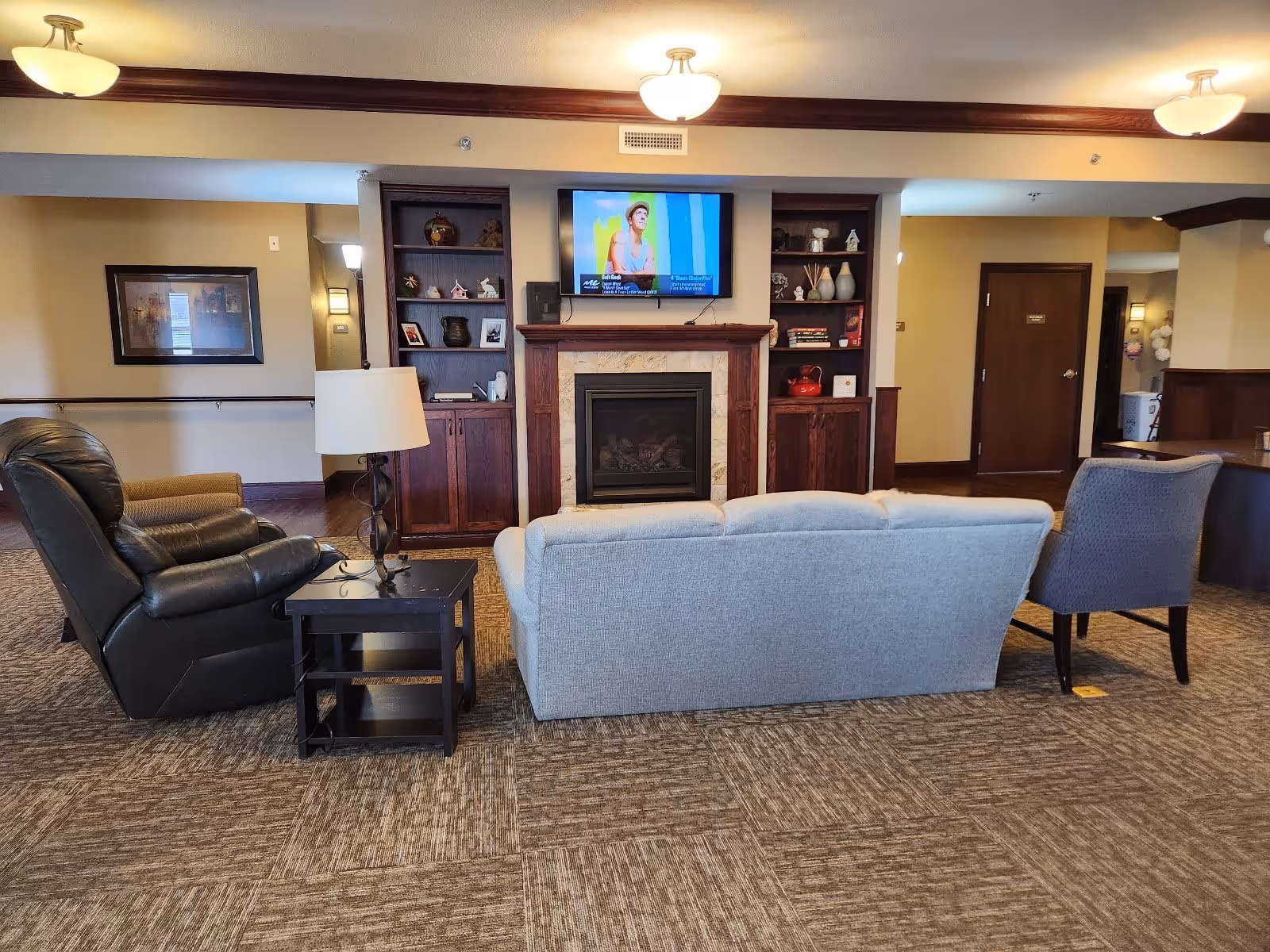 A cozy senior living common area featuring a gray couch, a black leather recliner, and a gray upholstered chair arranged around a black side table with a lamp. In front of the seating is a fireplace with a mounted TV above it, flanked by built-in wooden shelves with decorative items. The room has carpeted flooring and warm lighting from ceiling fixtures.