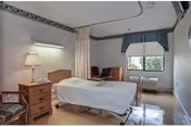 A simple, clean patient room in a skilled nursing facility featuring a single hospital bed with white linens, a wooden nightstand with a lamp and phone, a chair with striped upholstery, a small desk with a computer, and a window with a valance allowing natural light to enter.