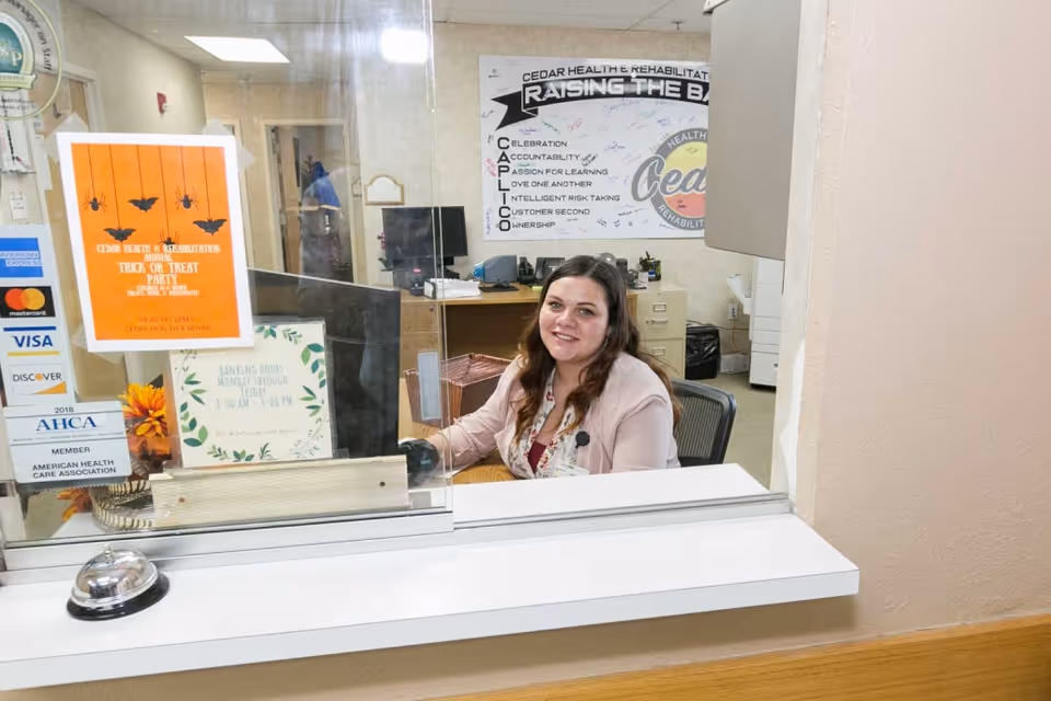 A receptionist smiles behind a protective glass window at the Cedar Health and Rehabilitation front desk with signs and a service bell.