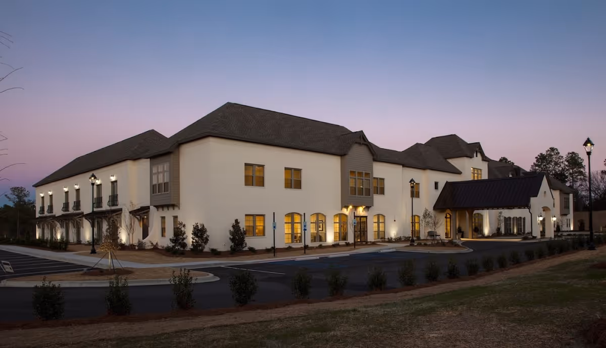 A modern building exterior of The Blake at Woodcreek Farms, featuring a combination of white walls and a dark roof, with warm lights illuminating the windows at dusk.