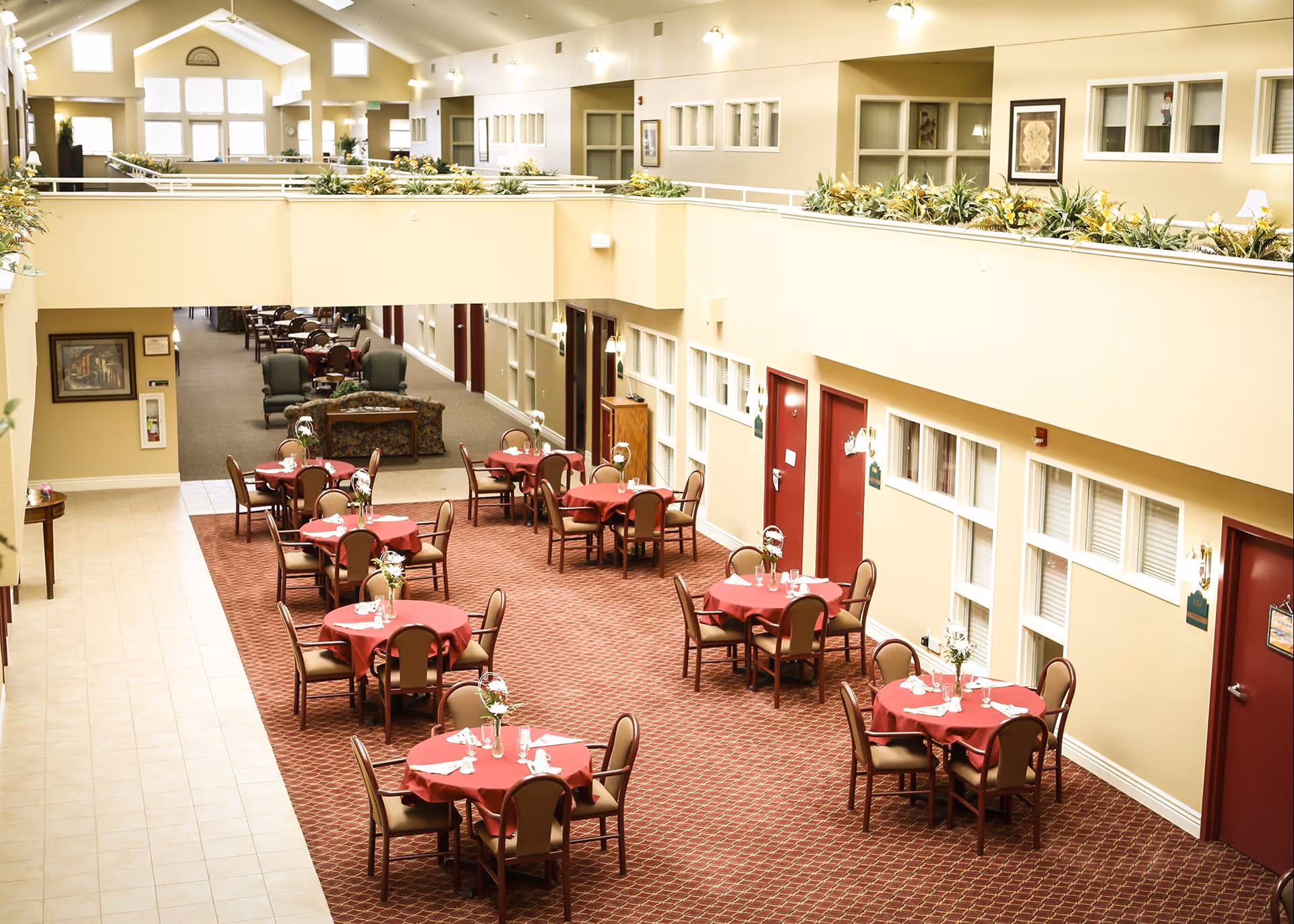 Interior view of a senior living facility dining area with multiple round tables covered with red tablecloths, each set with napkins and small flower arrangements. The space has beige walls, red doors, and a carpeted floor. There is a second level with plants along the railing and large windows letting in natural light.