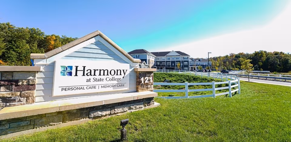 Entrance sign reading 'Harmony at State College' with the senior living building and landscaped grounds behind a white fence.