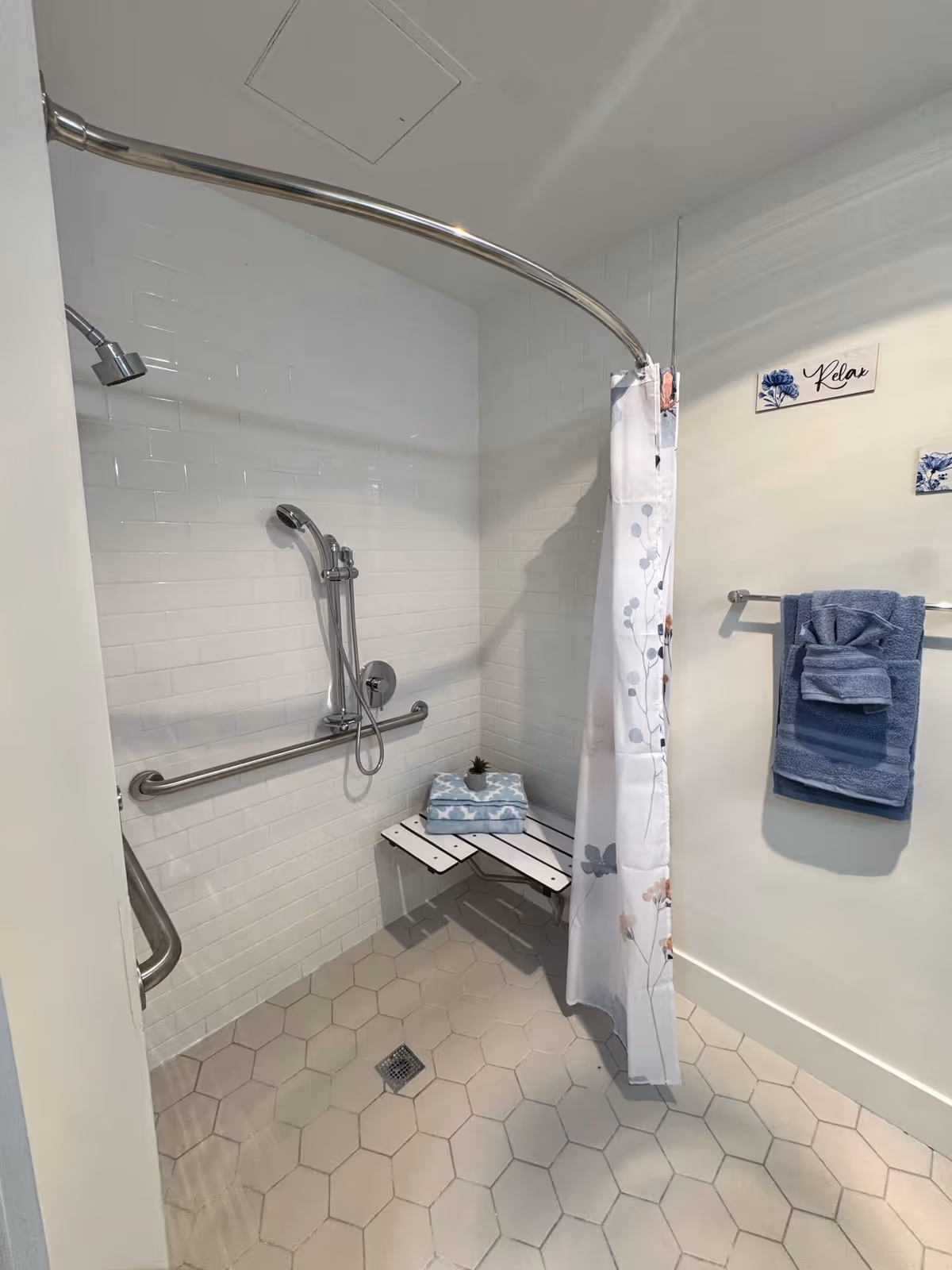 A bathroom shower area with white hexagonal floor tiles and white subway tile walls. The shower has a curved metal shower rod with a floral-patterned shower curtain partially drawn. There is a wall-mounted handheld showerhead and a horizontal grab bar. A fold-down white shower bench holds folded blue towels and a small potted plant. On the adjacent wall, a towel rack holds neatly folded blue towels, and decorative wall plaques with floral designs and the word 'Relax' are visible.