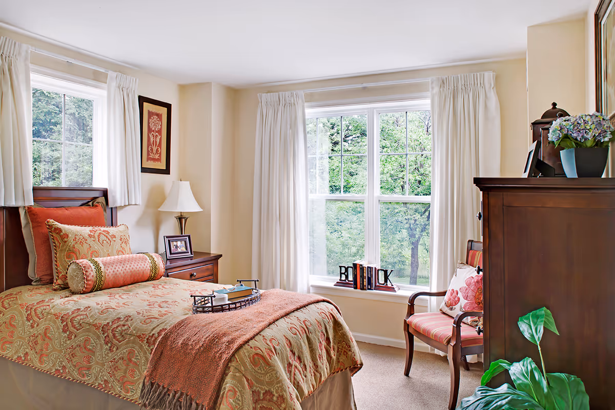 Sunlit furnished bedroom with a bed, nightstand, armchair, dresser, and a large window overlooking greenery.