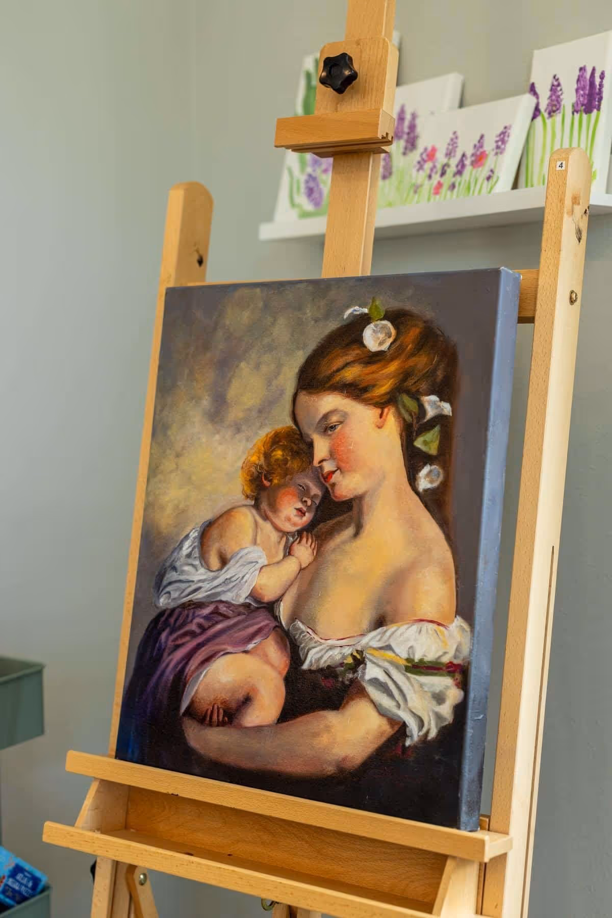 A painting on an easel depicting a woman holding a sleeping child close to her chest, with flowers in her hair and a soft, warm background. Behind the easel, there are paintings of purple flowers on a white shelf.