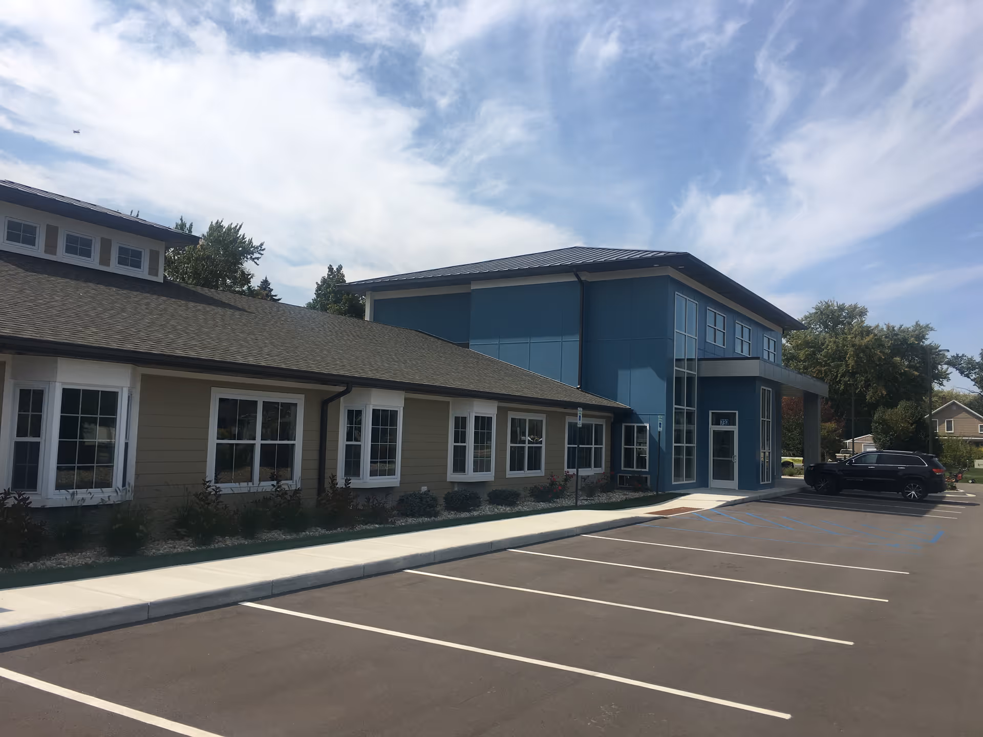 Exterior view of a modern two-story memory care building with a blue entrance and empty parking lot.