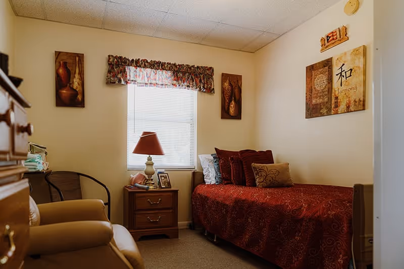A cozy bedroom in an assisted living facility featuring a single bed with red patterned bedding and multiple pillows, a wooden nightstand with a lamp and framed photo, a window with floral valance, two framed artworks on the walls, and a comfortable armchair in the foreground.