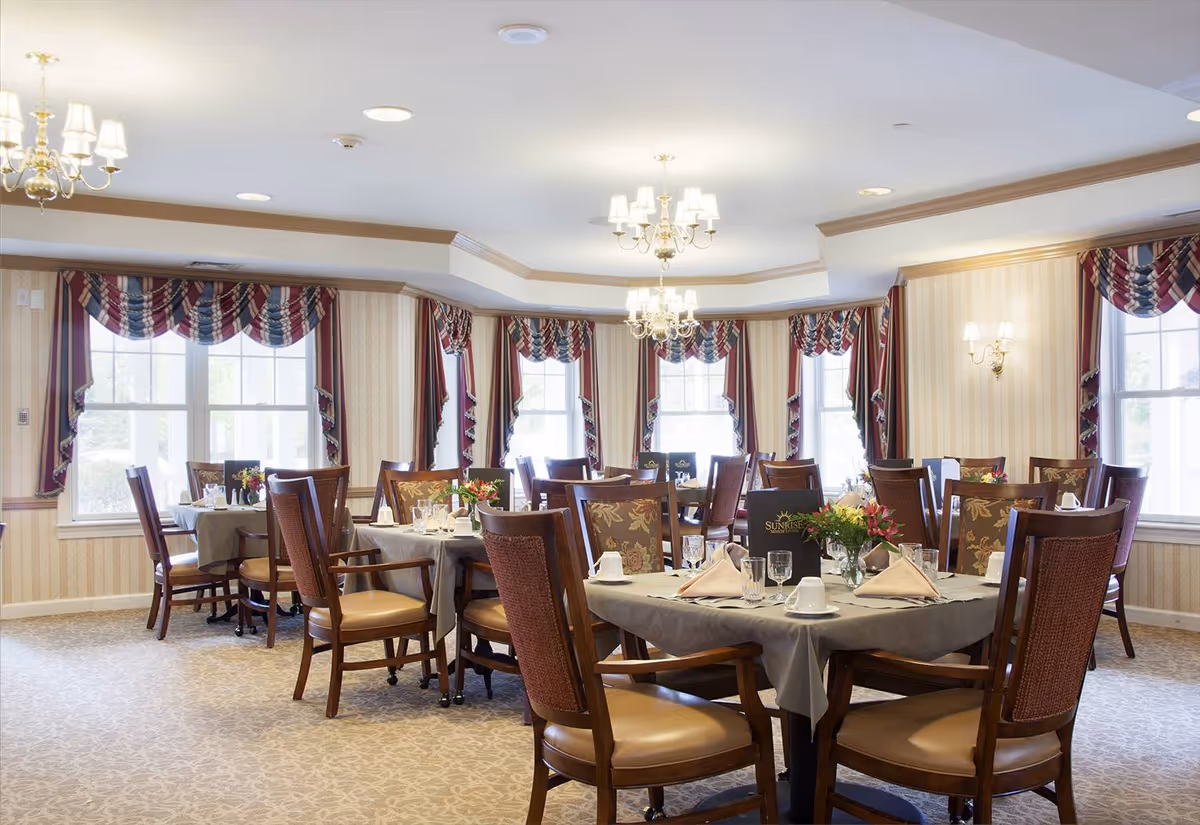 A bright and elegant dining room with multiple round tables covered with gray tablecloths, set with white cups, glasses, napkins, and floral centerpieces. The room features large windows with red, white, and blue valances, beige patterned wallpaper, and carpeted floors. Chandeliers and wall sconces provide warm lighting.