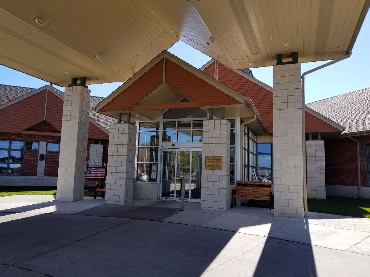 Entrance of a building with a covered drop-off area supported by large square columns. The building has a peaked roof with red siding and large glass windows around the entrance door. There are benches on either side of the entrance and a plaque mounted on one of the columns.