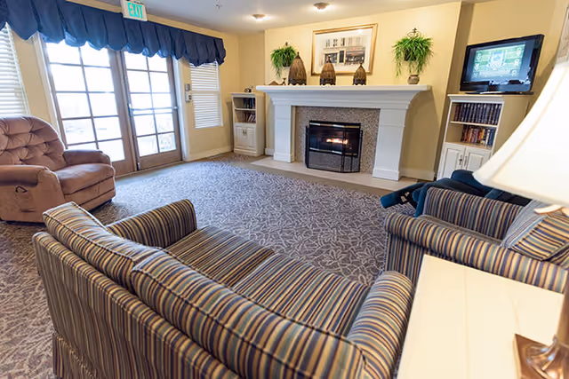 A cozy senior living lounge with striped sofas and armchairs facing a fireplace, TV, and glass French doors.