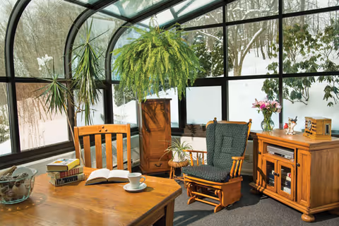 A sunroom with large curved glass windows showing a snowy outdoor scene. Inside, there is a wooden table with an open book, a cup and saucer, and a stack of books. A wooden chair and a cushioned rocking chair are positioned near the table. There are several plants, including a large hanging fern and a tall potted plant. A wooden cabinet with a vase of flowers and decorative items is also visible.