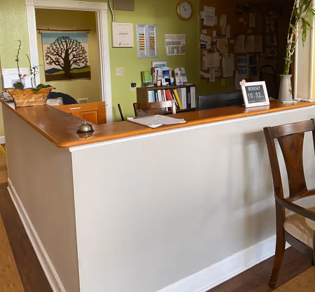 Reception desk area in a green-walled interior with a service bell, chairs, bulletin board, clock, and decorative wall hanging.