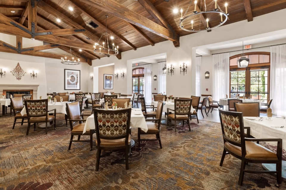 A spacious dining room with multiple tables covered in white tablecloths, each set with napkins and small flower arrangements. The room features wooden ceiling beams, chandeliers, wall sconces, large windows with sheer curtains, and a fireplace on one side. The chairs have patterned backs and leather seats.