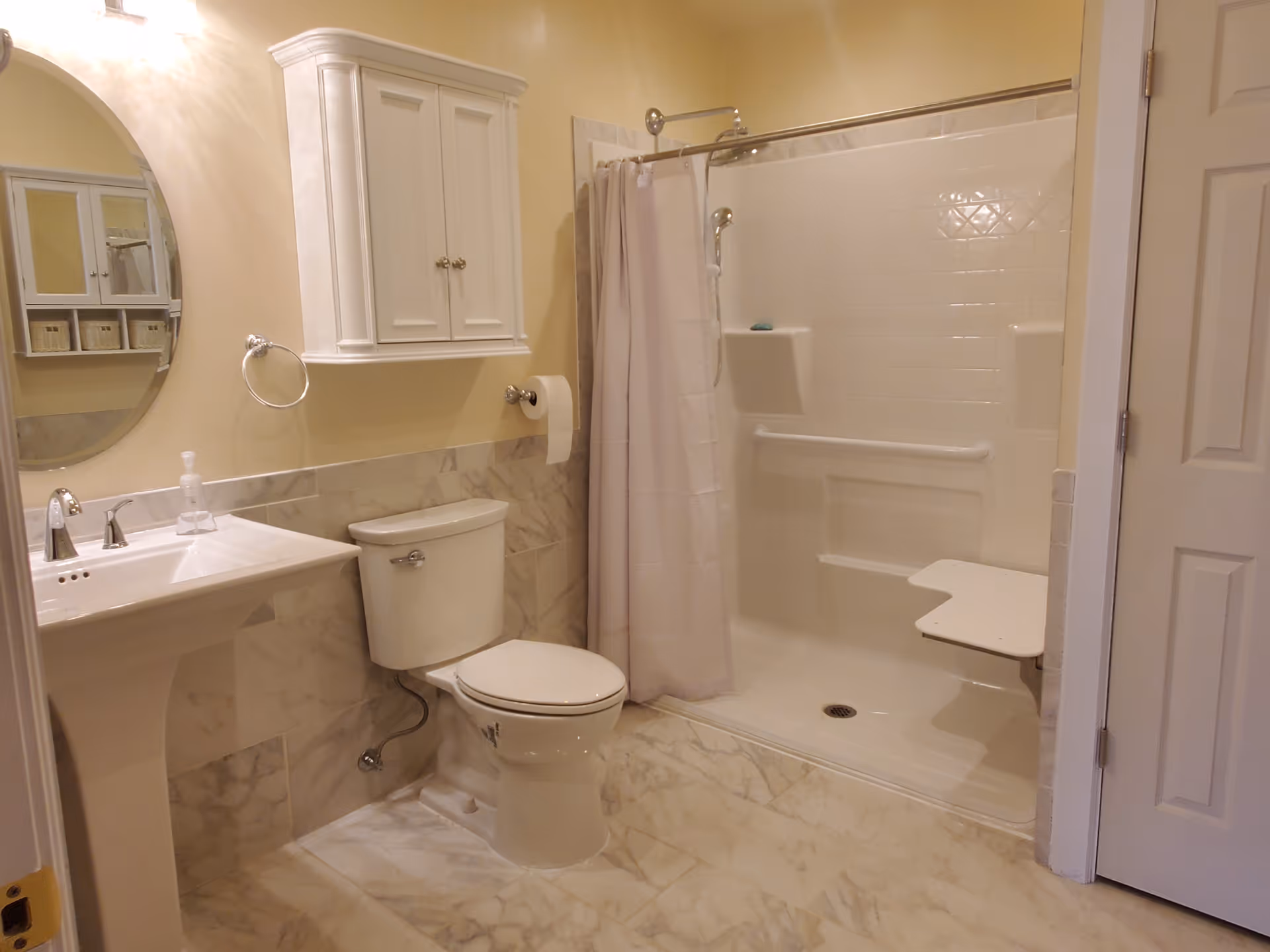 A clean bathroom with a white pedestal sink, a toilet, a wall-mounted cabinet, a round mirror, and a walk-in shower with a curtain and a built-in foldable seat.