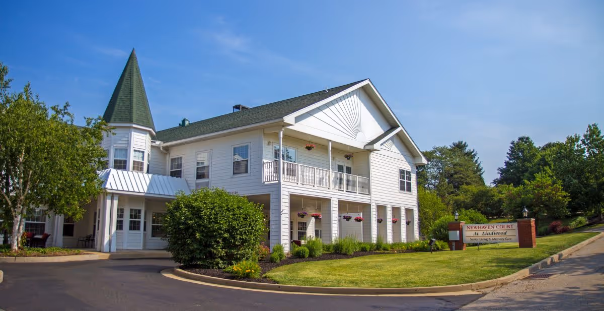 Exterior view of Newhaven Court at Lindwood, a two-story white building with a green roof and a pointed turret. The building is surrounded by well-maintained landscaping including bushes, trees, and hanging flower baskets. A sign near the driveway entrance reads 'Newhaven Court at Lindwood Senior Living & Memory Care'. The sky is clear and blue.