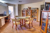 Bright common room with a round table and four chairs, bookshelves, a desk and a sliding glass door leading to a balcony.