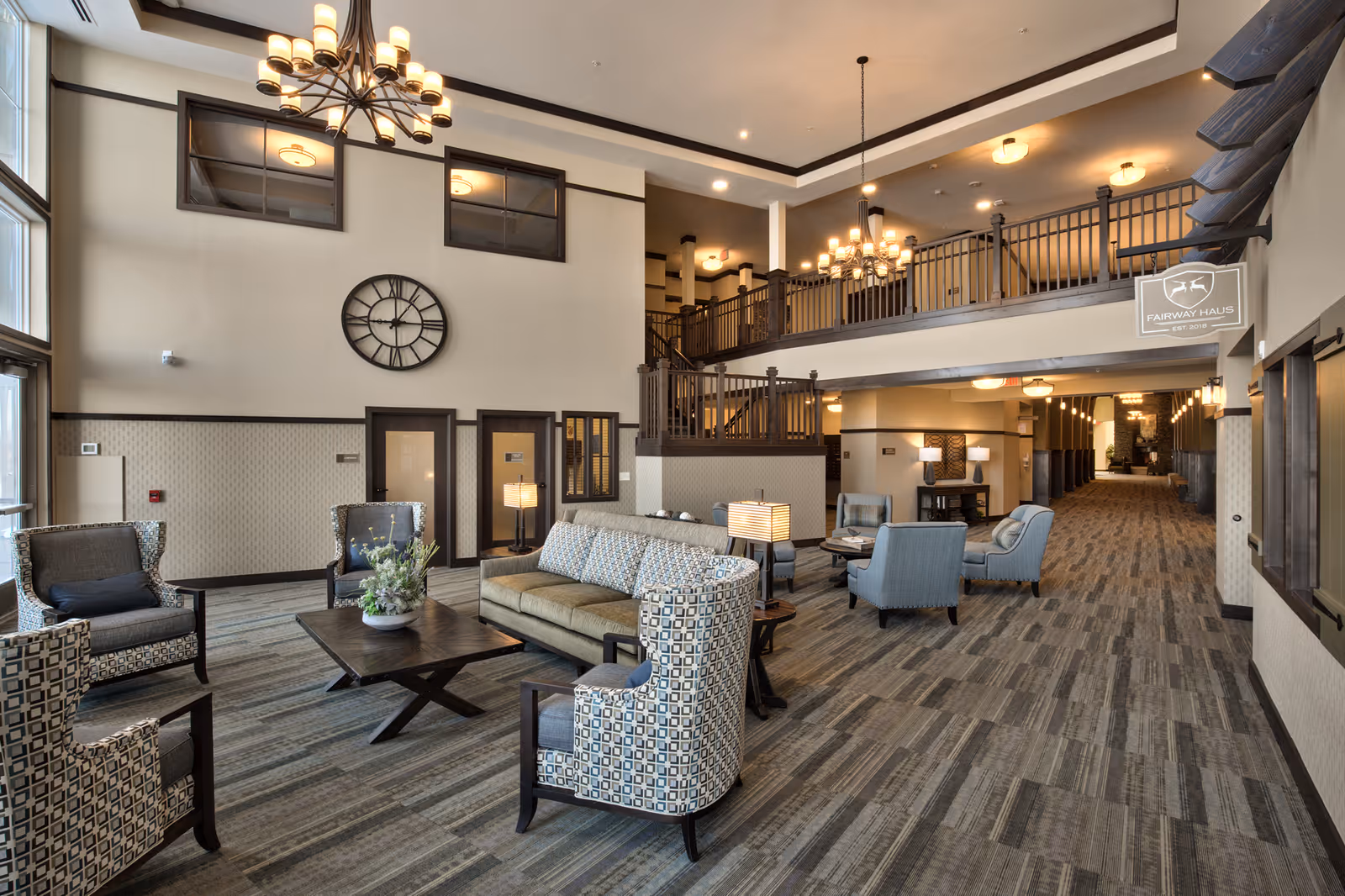 Spacious and well-lit senior living facility common area with high ceilings, large windows, and a mezzanine level. The room features comfortable seating including patterned armchairs and a sofa around a wooden coffee table with a floral centerpiece. There are multiple chandeliers and lamps providing warm lighting, a large wall clock, and a long carpeted hallway extending into the background.