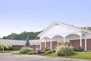 Front entrance of a single-story brick healthcare and rehabilitation center with a white covered portico, landscaped shrubs, and a driveway.