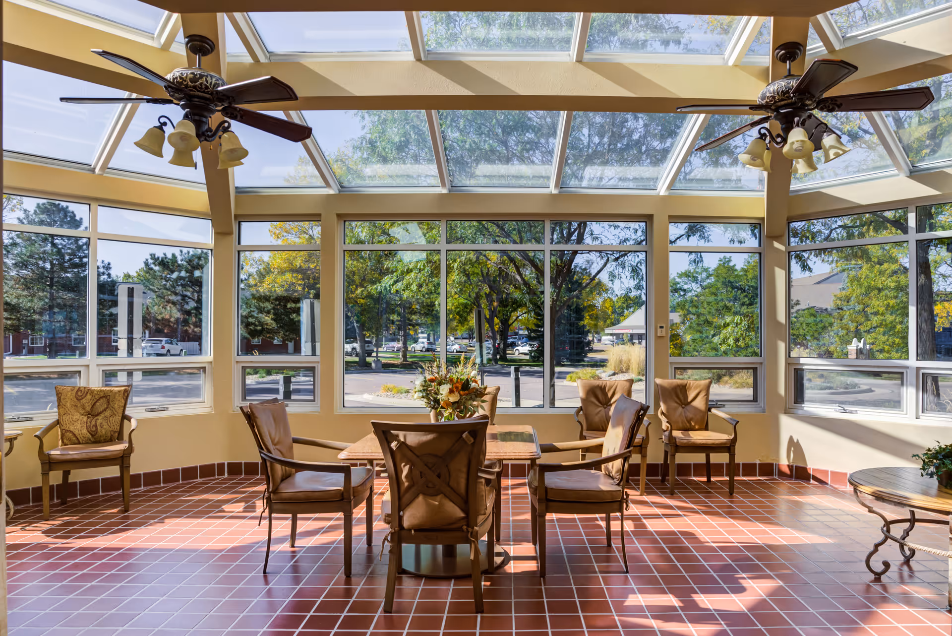A bright sunroom with large windows and a glass ceiling, furnished with a table surrounded by six cushioned chairs. Two ceiling fans with lights hang from the ceiling. Outside, trees and a parking lot are visible.