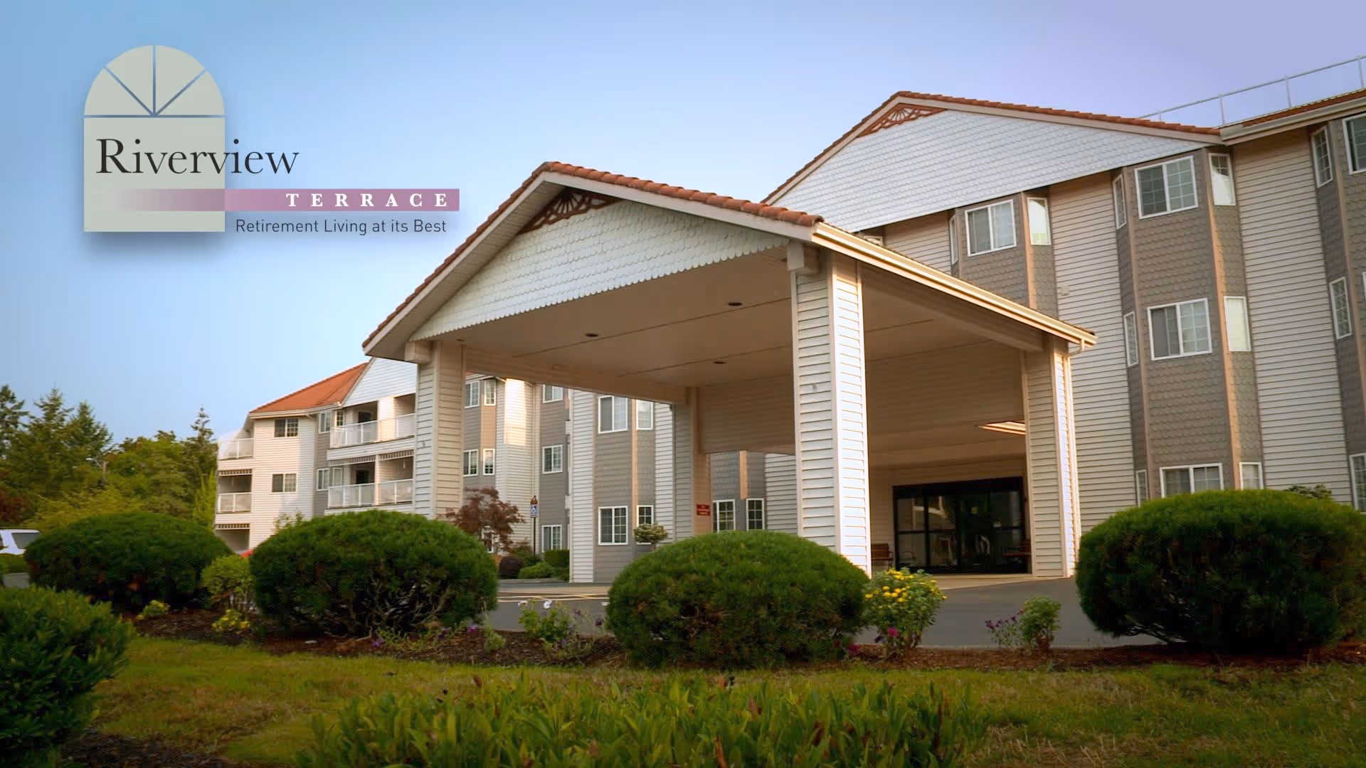Exterior view of Riverview Terrace, a multi-story retirement living facility with a covered entrance, surrounded by green bushes and trees under a clear sky.