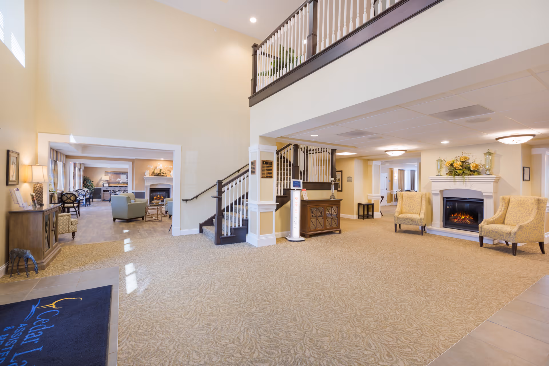 Spacious and well-lit interior common area of Cedar Lake Assisted Living and Memory Care featuring a staircase with white railings and dark wood banister, a cozy seating area with two yellow armchairs by a fireplace adorned with flowers, and an adjacent lounge area with additional seating and a second fireplace. The walls are painted in warm beige tones and the carpet has a subtle pattern. A dark blue mat with the facility's name is visible near the entrance.