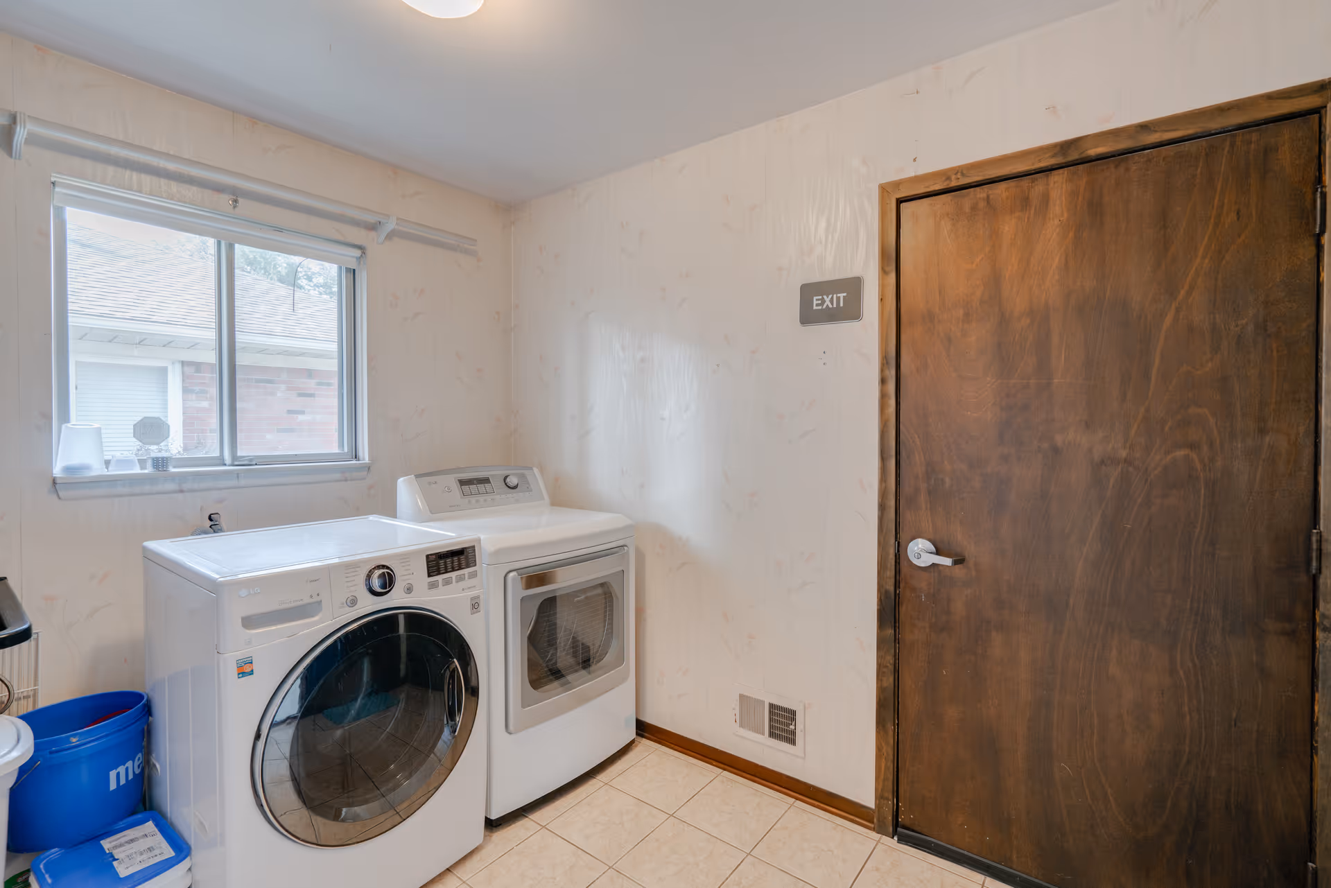 Laundry room with a front-loading washing machine and a dryer side by side under a window. There is a wooden door with an exit sign on the wall above it. The floor is tiled and the walls have a light floral wallpaper.