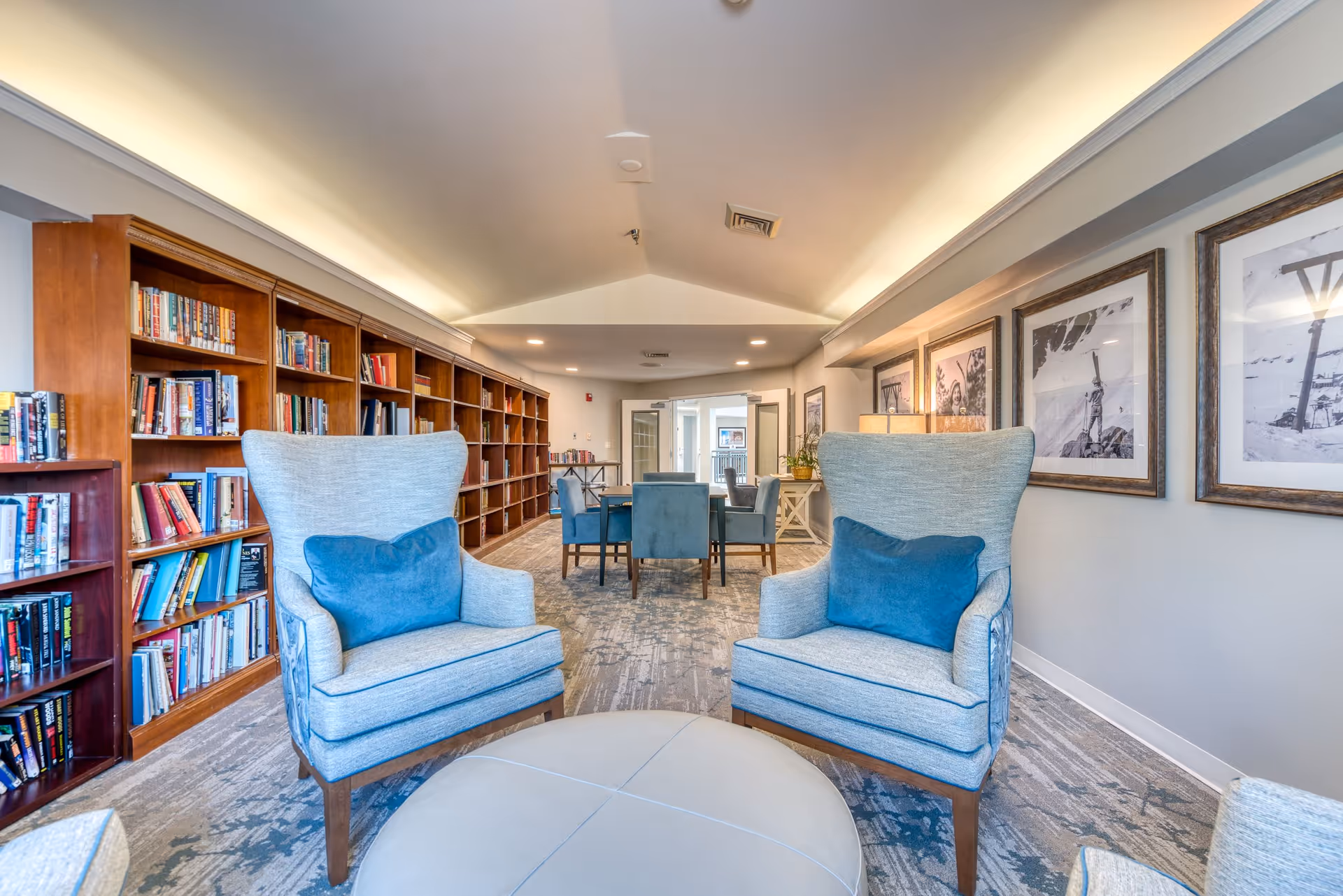 A cozy senior living common area with two light blue armchairs with blue cushions facing a round ottoman. Behind the chairs is a long wooden bookshelf filled with books along the left wall. In the background, there is a table with six blue upholstered chairs. The right wall features framed black and white photographs and a lamp on a side table. The room has a carpeted floor and a high ceiling with recessed lighting.